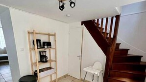 A spacious room with a tiled floor, a wooden ladder shelf, a black trash bin, a white chair, and a wooden staircase.