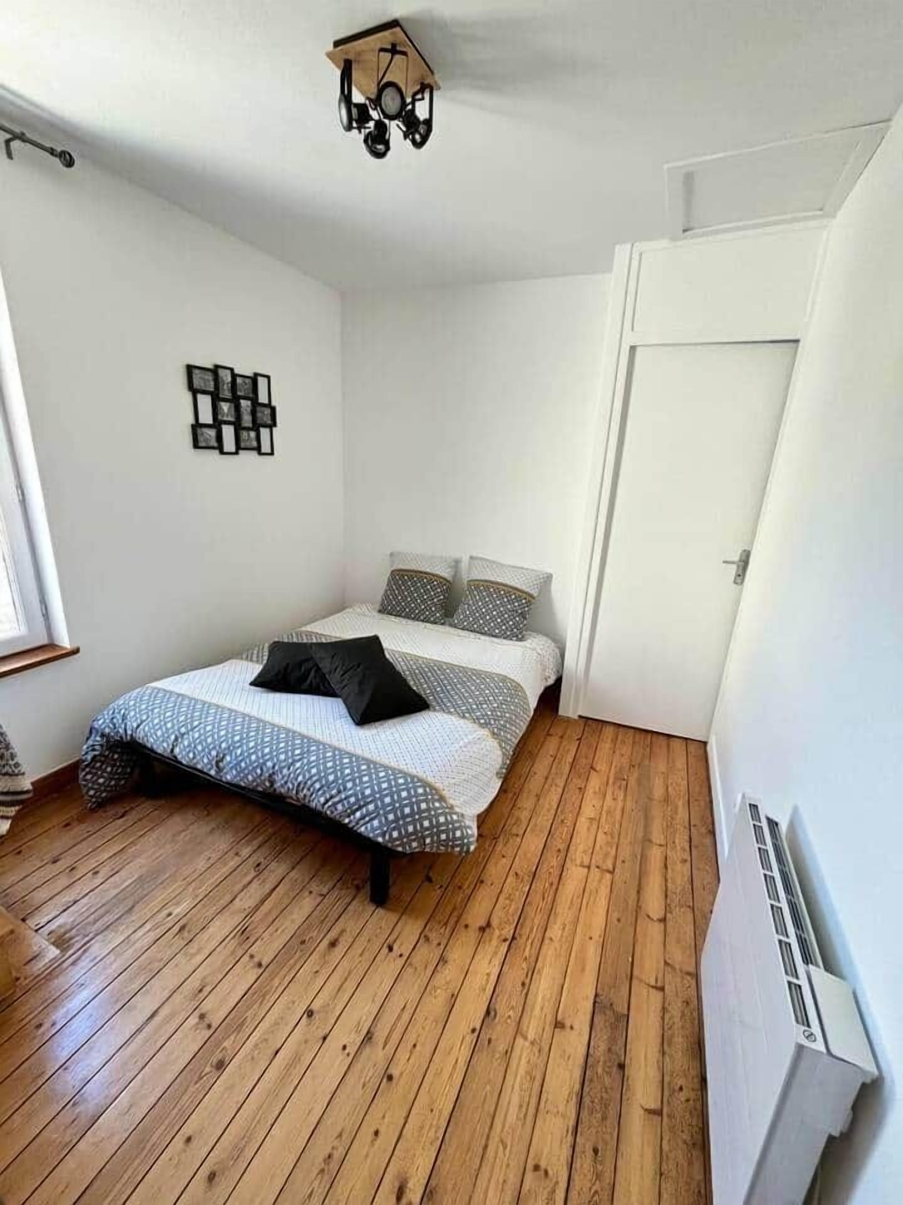 A bedroom with a wooden floor, a bed with patterned bedding, a wall-mounted light fixture, and a white door.