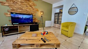 A living room with a wooden coffee table, a TV on a wooden stand, a yellow armchair, and a bookshelf.