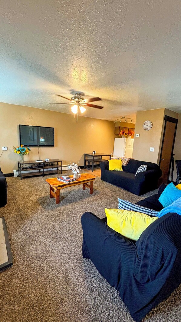 A living room with a ceiling fan, a flat-screen TV, a coffee table, a sofa with cushions, and a small table with books and a vase.