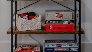 Une bibliothèque en bois, sur l’étagère supérieure de laquelle on peut lire le mot « VINO », ainsi qu’une horloge et divers jeux de société et livres répartis sur les autres étagères.