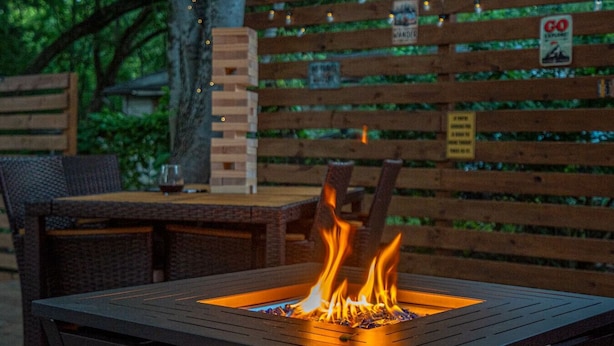 Un foyer carré avec des flammes, une clôture en bois ornée de panneaux et du mobilier d’extérieur.