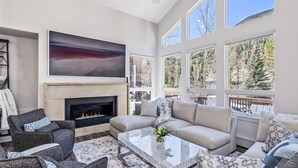 A modern living room with a fireplace, large windows, and a view of trees. - 4 Bedroom Luxury Home in East Vail (Vail)