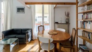 Ett vardagsrum med en svart soffa, ett soffbord i glas, ett matbord i trä med stolar, en bokhylla och en utsikt genom ett fönster.