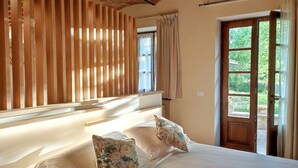 Une chambre avec une tête de lit en lattes de bois, une grande fenêtre avec des rideaux et une vue sur la verdure à l’extérieur.