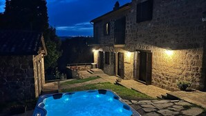 Un bâtiment en pierre avec un jacuzzi en premier plan, un chemin en pierre et une vue sur le paysage environnant au crépuscule.