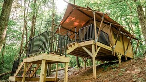 Een boomhut met een houten terras en metalen leuning, omgeven door een bos.