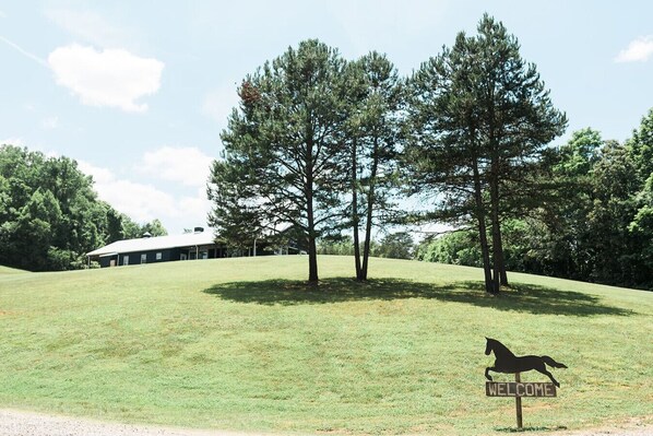 A grassy area with a welcome sign and a horse silhouette, surrounded by trees and a building in the background.