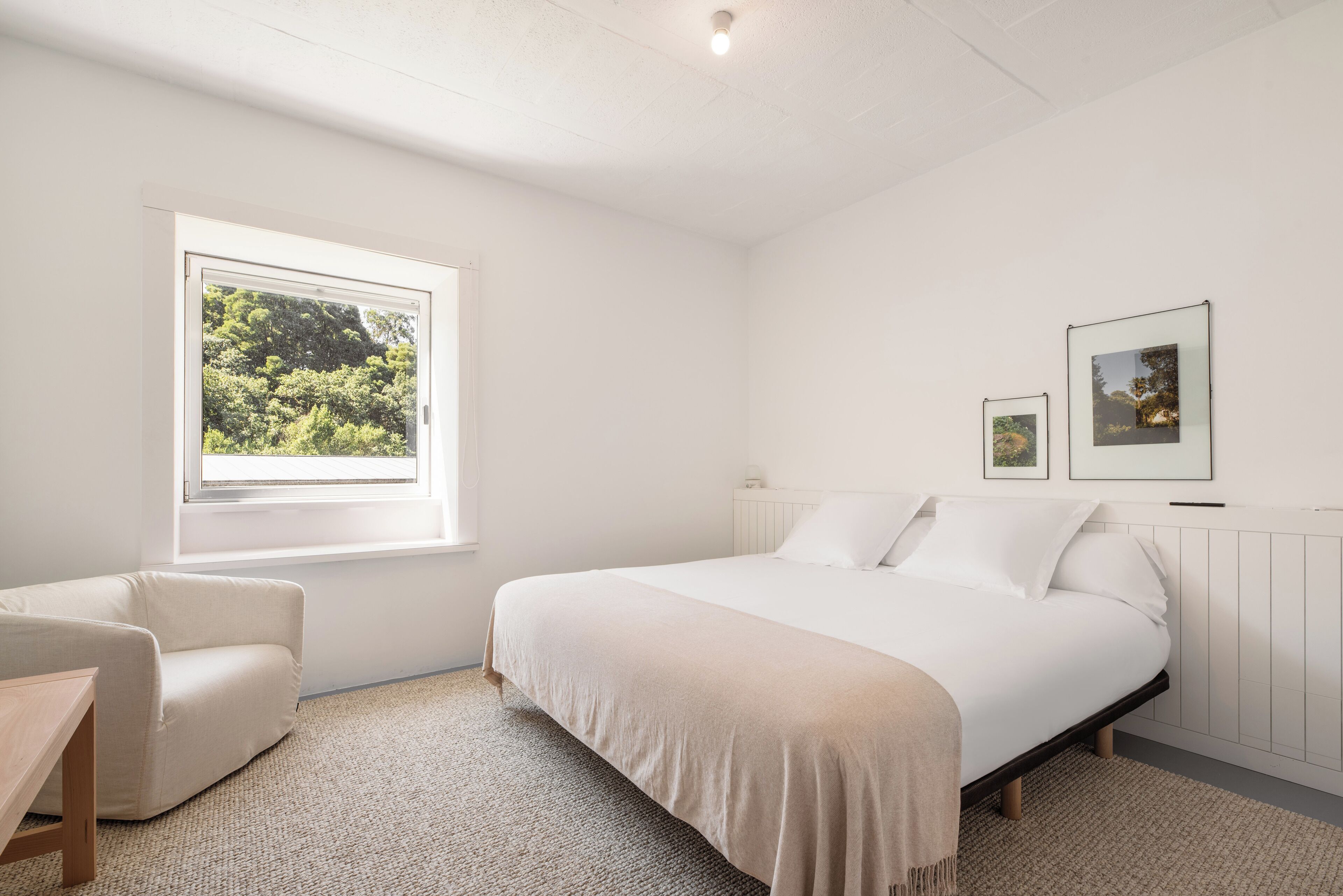 A bedroom with a large bed, a window with a view of greenery, a chair, and two framed pictures on the wall.