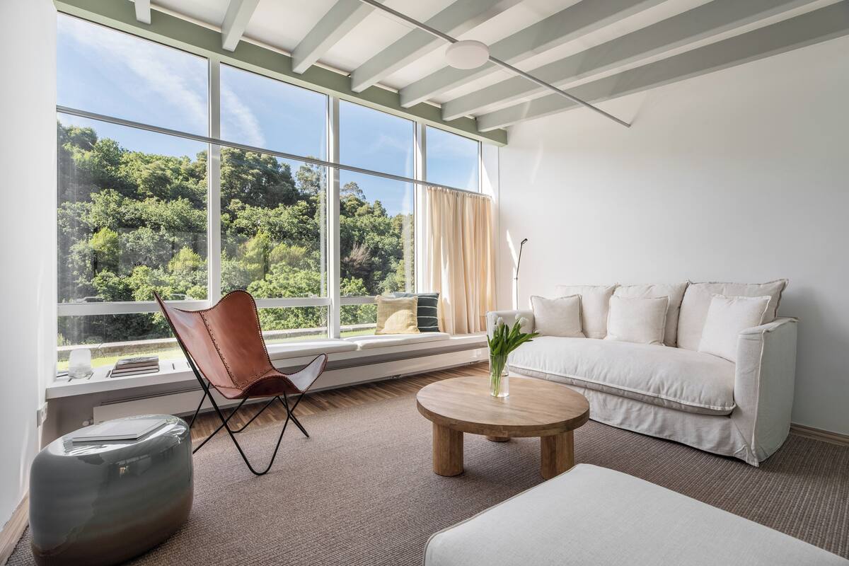 Un salon moderne avec une grande fenêtre, un canapé blanc, une table basse en bois, un fauteuil en cuir et un pouf en verre.