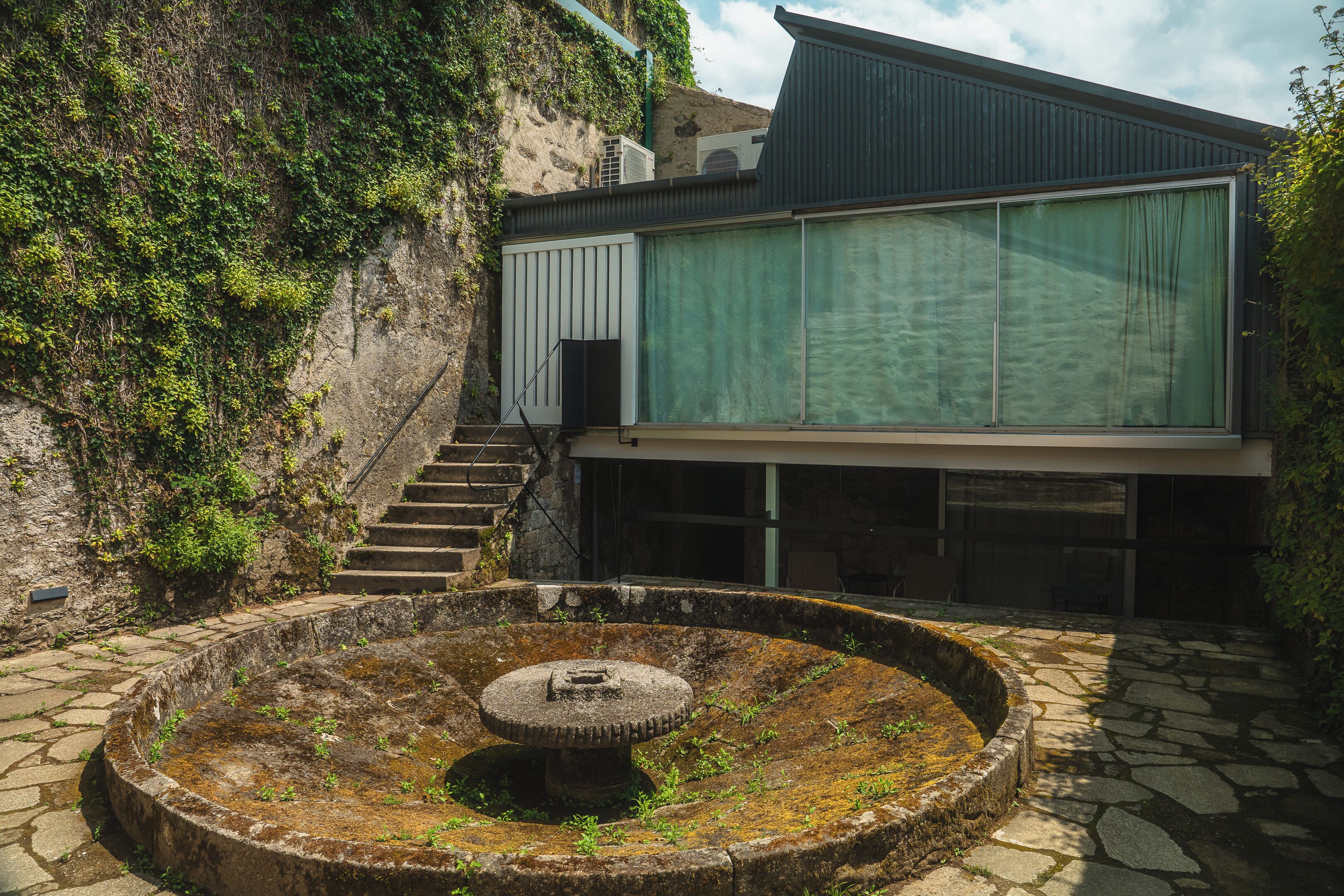 A building with a stone staircase leading to a modern entrance, a circular stone fountain, and a view of a hillside with vegetation.