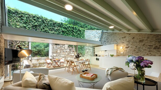 A modern living room with a sofa, a dining area with a table and chairs, a kitchen area, and a large window overlooking greenery.