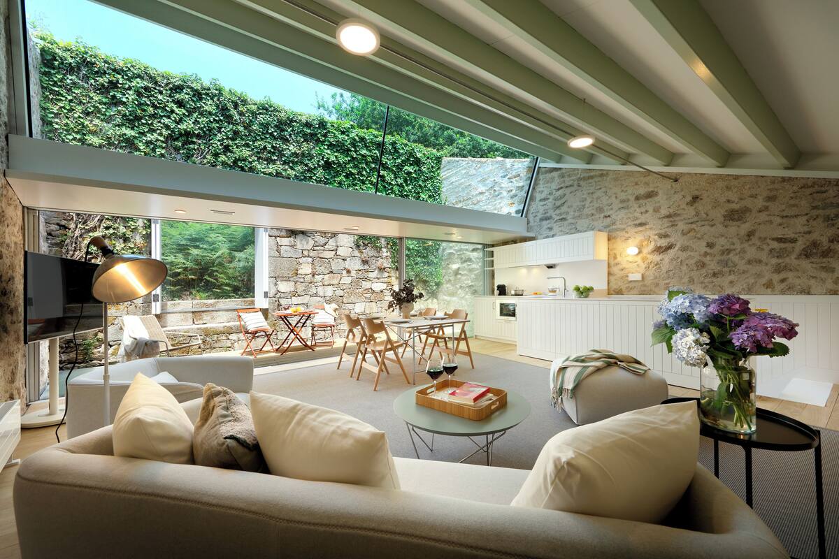 Un salon moderne avec un canapé, un coin repas comprenant une table et des chaises, un espace cuisine et une grande fenêtre donnant sur un espace verdoyant.