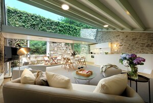 Una sala de estar moderna con un sofá, un comedor con mesa y sillas, una zona de cocina y un ventanal con vistas a zonas verdes.