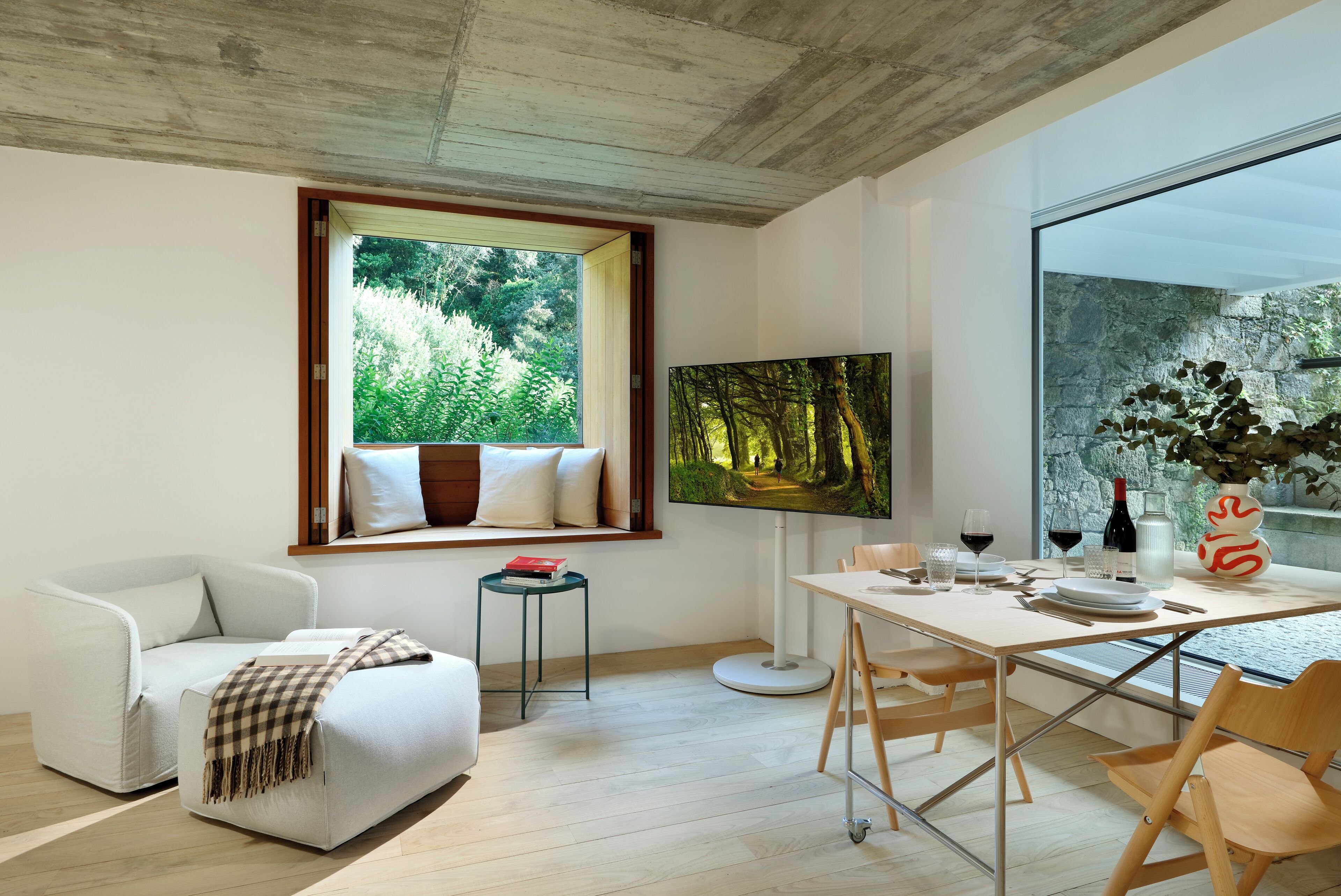 A modern living room with a dining area, a large window with a view of greenery, a flat-screen TV, and a dining table set with glasses and a vase.