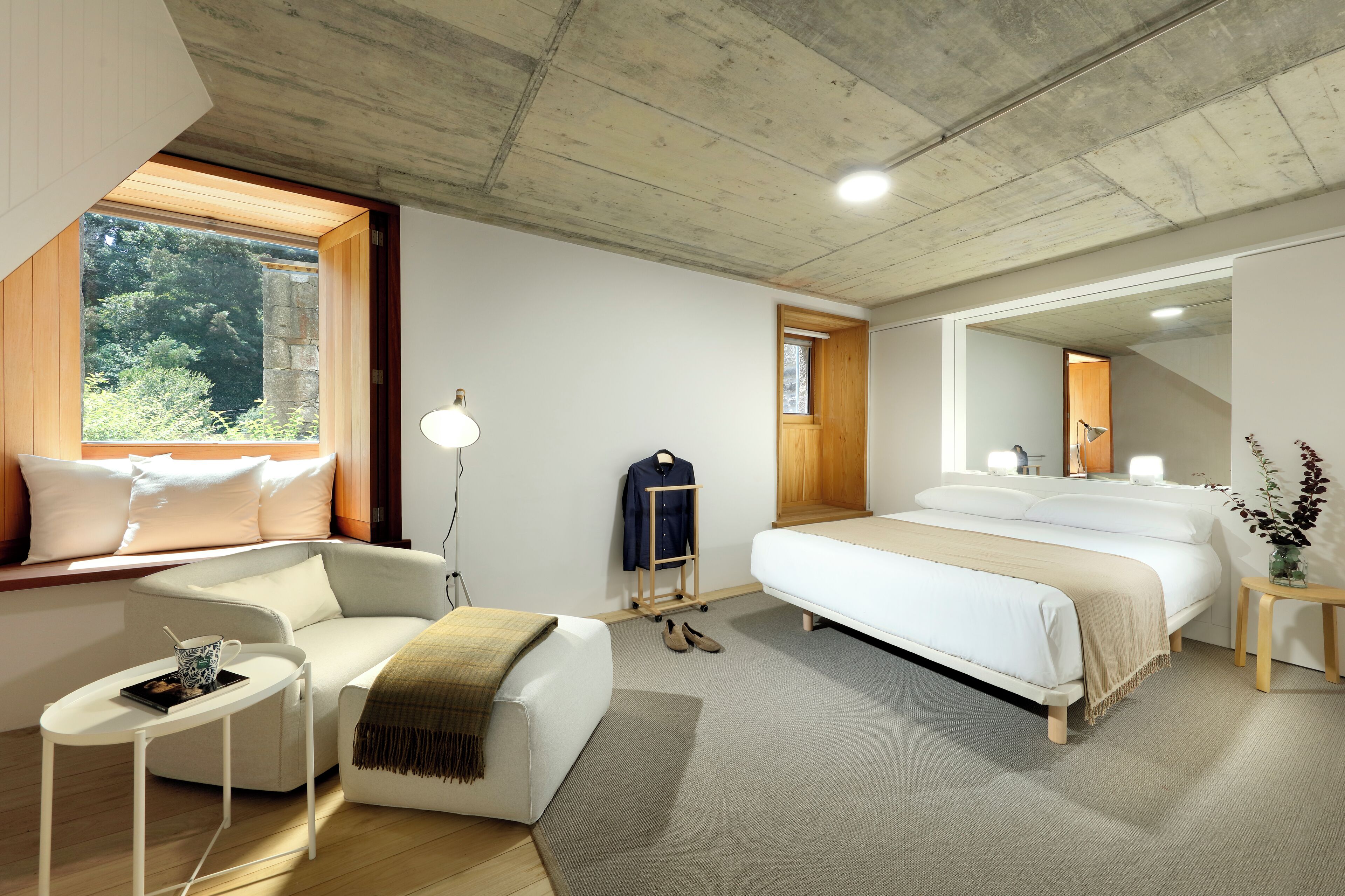 A modern bedroom with a large bed, a sitting area with a small table, a wardrobe, and a window with a view of greenery.