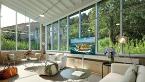A modern living room with large windows, a flat-screen TV, comfortable seating, and a view of greenery.