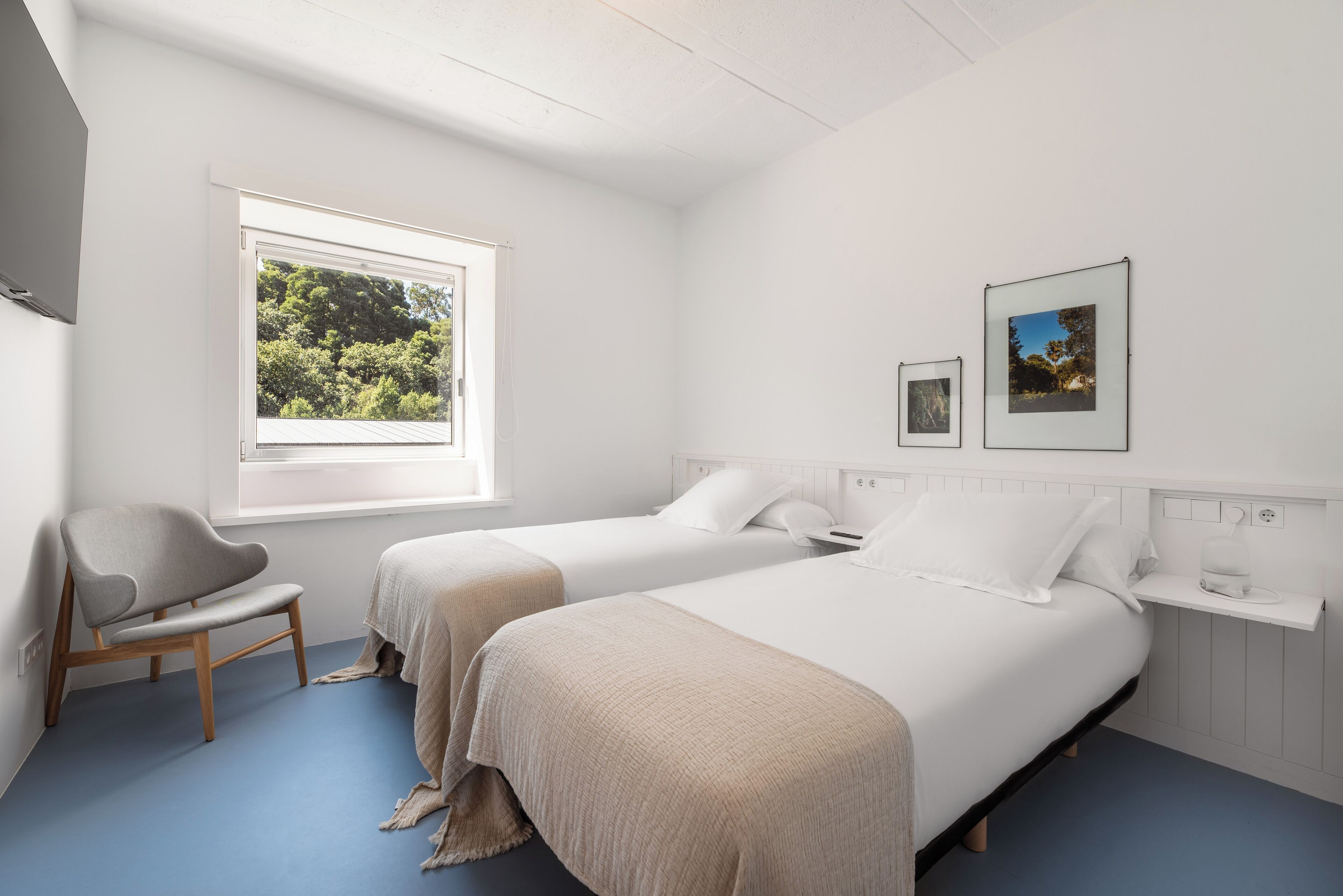 A bedroom with two beds, a window with a view of trees, a chair, and a bedside table with a lamp.