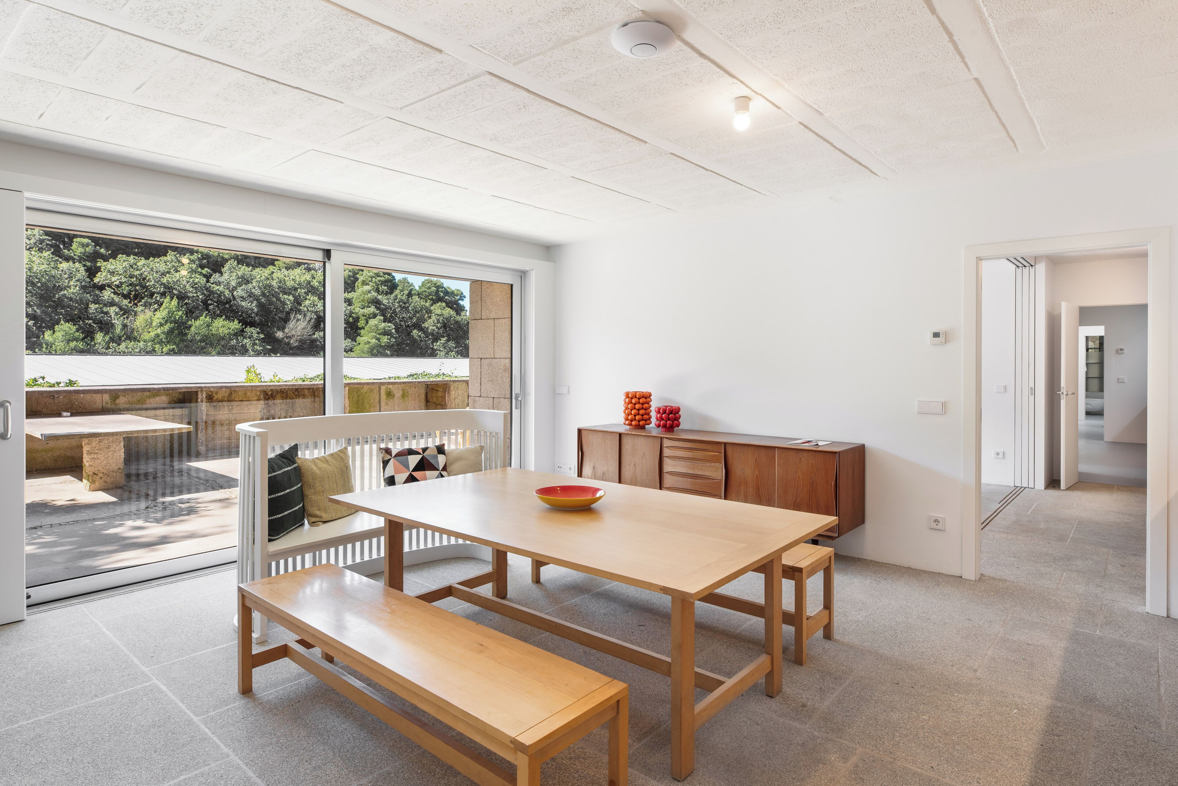 Un comedor moderno con una mesa y banco de madera, un vidrio que da a una zona de estar al aire libre, y vistas de vegetación.