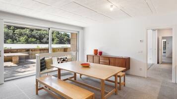 Un comedor moderno con una mesa y banco de madera, un vidrio que da a una zona de estar al aire libre, y vistas de vegetación.