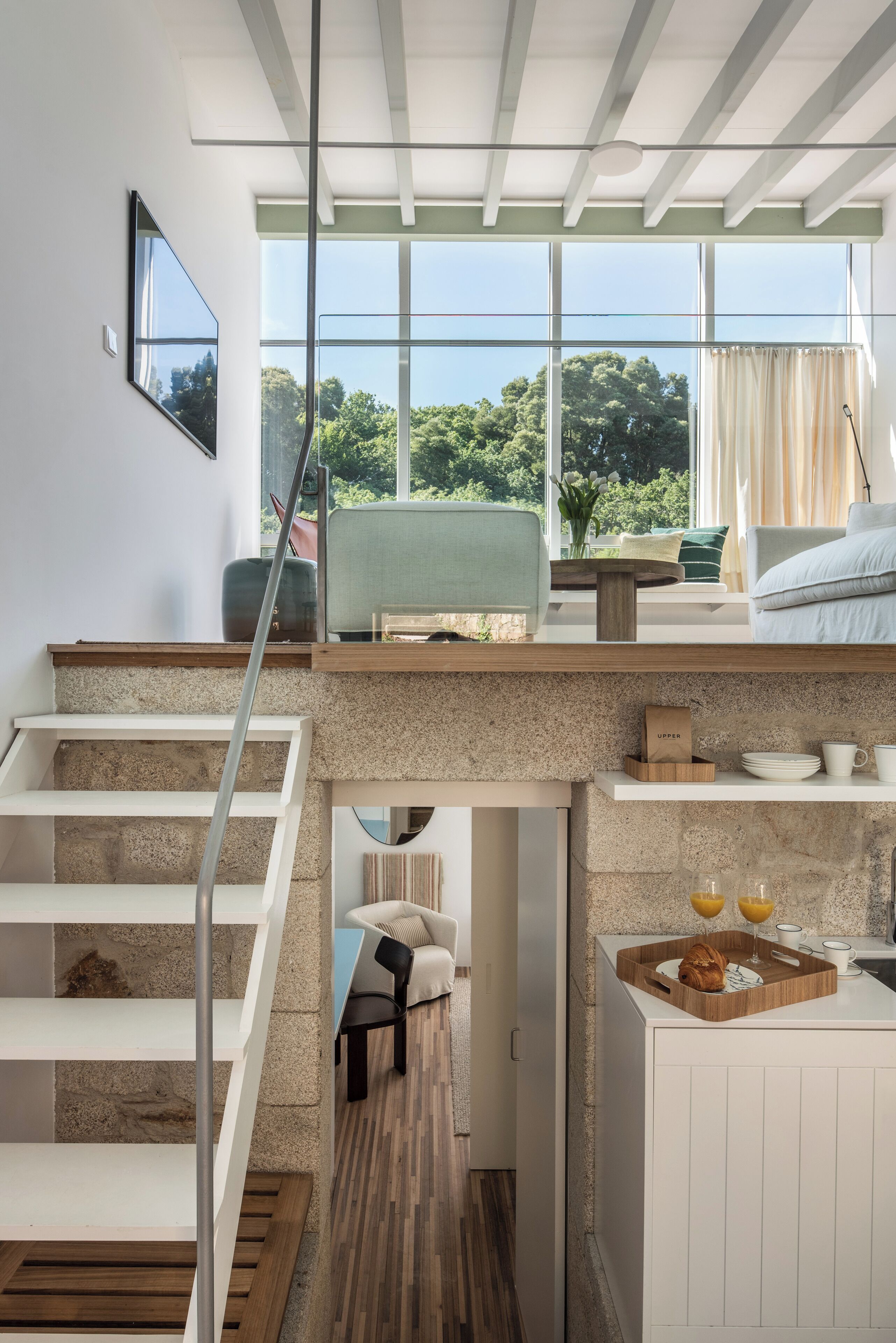 Een modern interieur met een trap naar een slaapkamer op een entresol, een keukenhoek met ontbijtartikelen, en uitzicht op groen buiten.