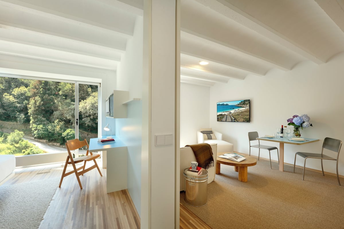 Une pièce moderne et bien éclairée, dotée d’une grande fenêtre, d’un bureau, d’une chaise, d’une petite table et d’un coin repas avec des chaises et une table.