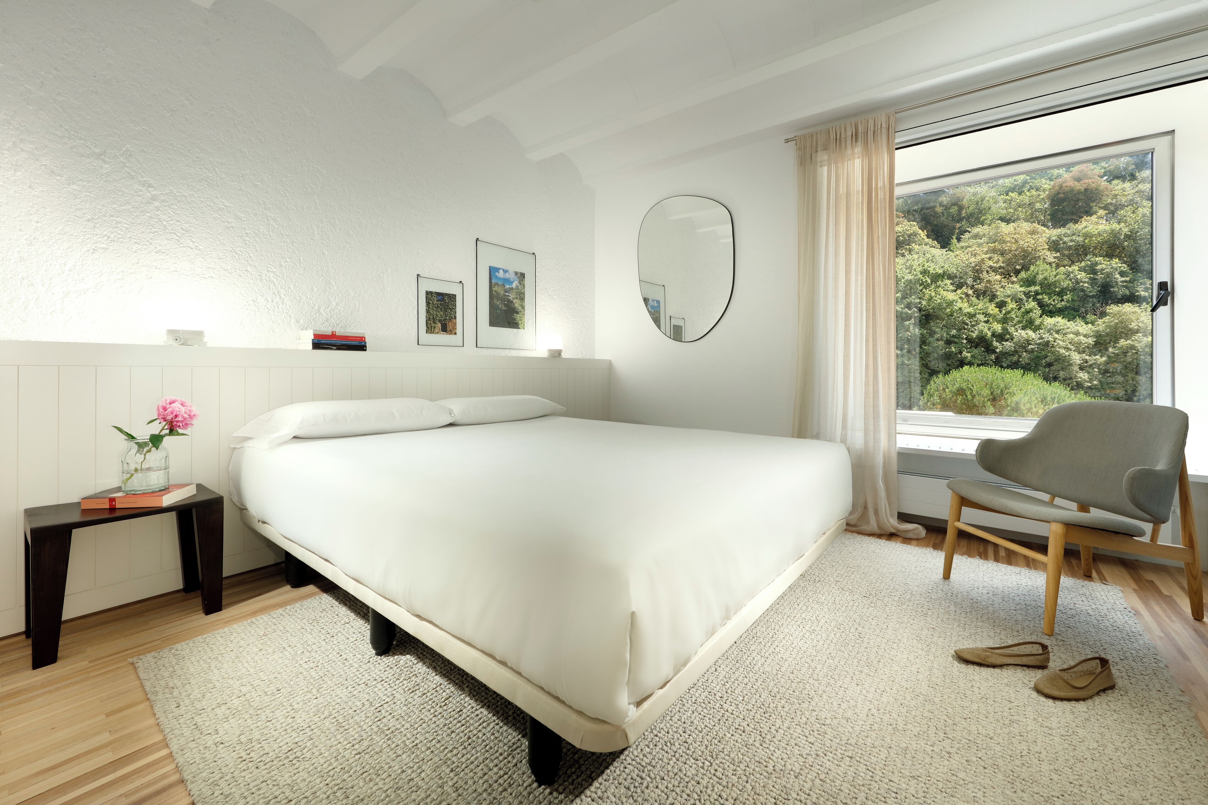 A bedroom with a large bed, a small table with a vase and books, a chair, and a view of trees through a window.