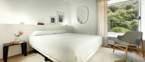 A bedroom with a large bed, a small table with a vase and books, a chair, and a view of trees through a window.