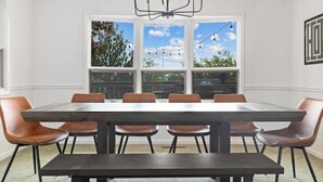 A dining area with a rectangular table and six chairs, a chandelier, and a large window with a view of trees and a clear sky.