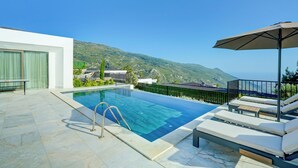 A swimming pool with lounge chairs and an umbrella, surrounded by a modern building and a view of mountains. - Pluto-1 PLANET LUXURY VILLAS (Kas)