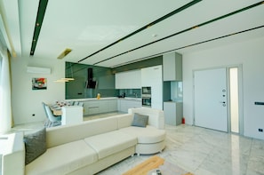 A modern living room with a white sectional sofa, a dining area with a table and chairs, and a kitchen with white cabinets and a large island.