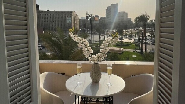 Ein Balkon mit zwei weißen Stühlen, einem kleinen Tisch mit einer Vase voller weißer Blumen und zwei Sektgläsern. In der Ferne sind Gebäude und Palmen zu sehen, die Sonne scheint.