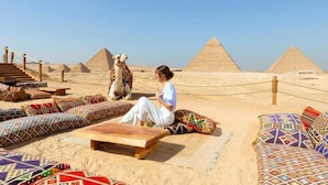 Eine Person sitzt auf einem gemusterten Kissen, im Hintergrund eine Kamel vor Pyramiden.