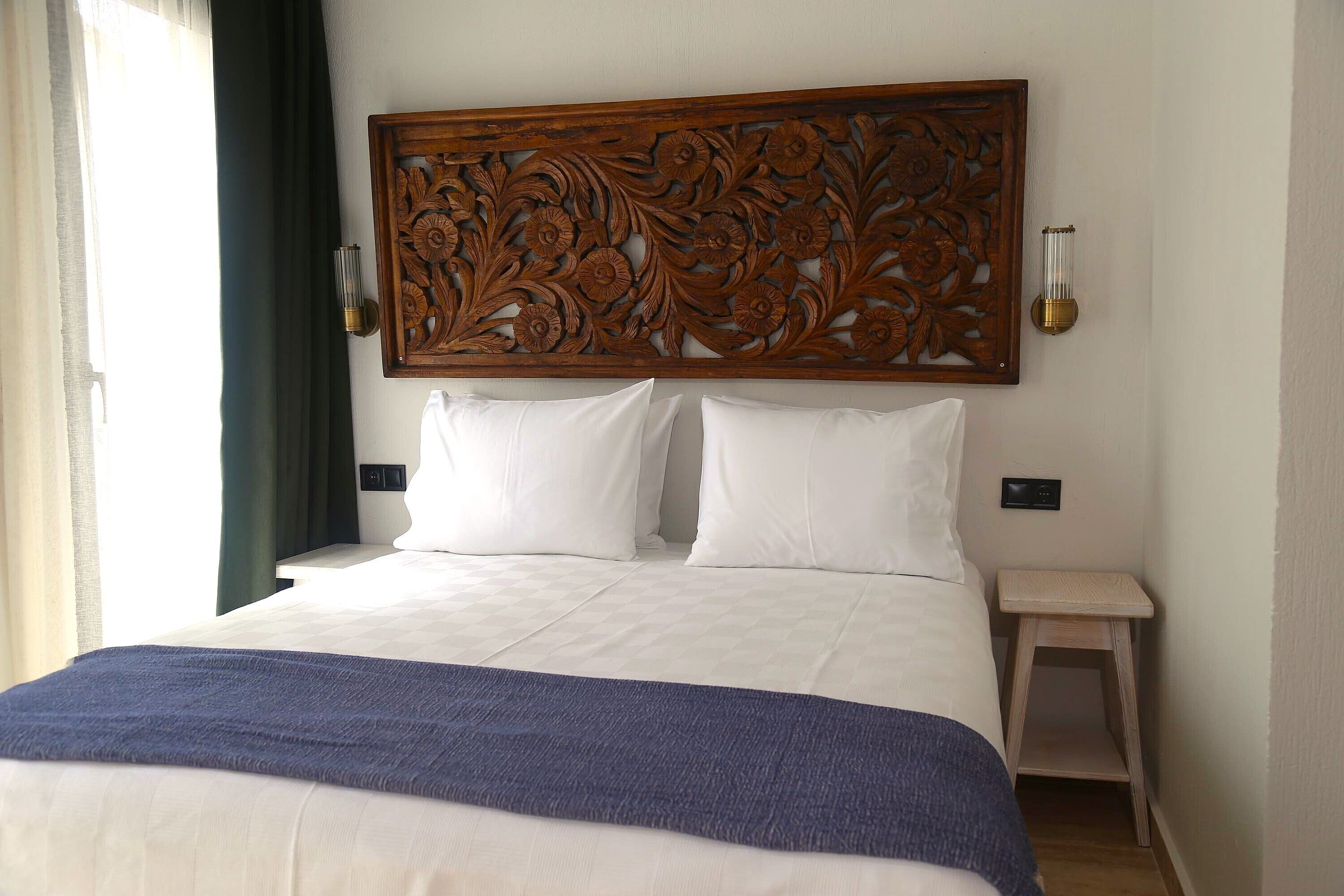 A neatly made bed with a blue blanket, a wooden headboard with floral carvings, and a small bedside table with a lamp.