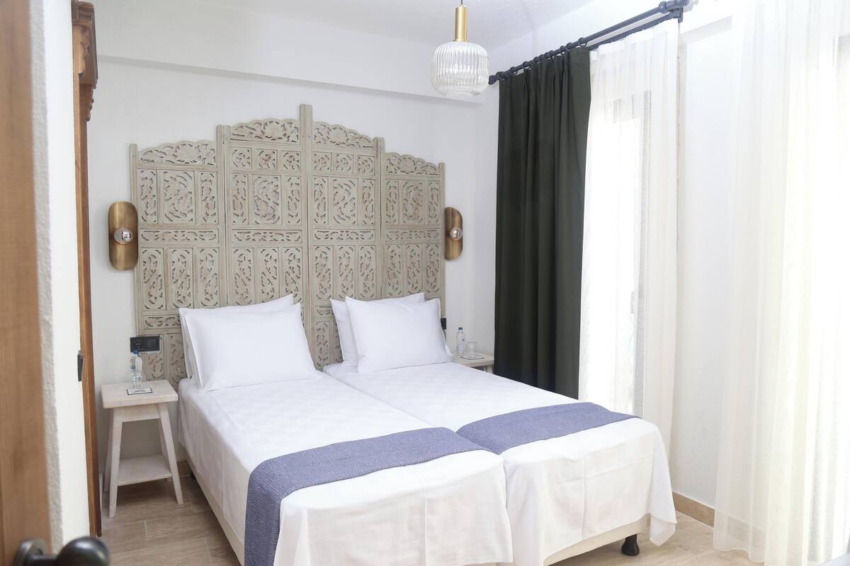 A double bed with a decorative headboard, a bedside table with a lamp, and a view of the outdoors through sheer curtains.