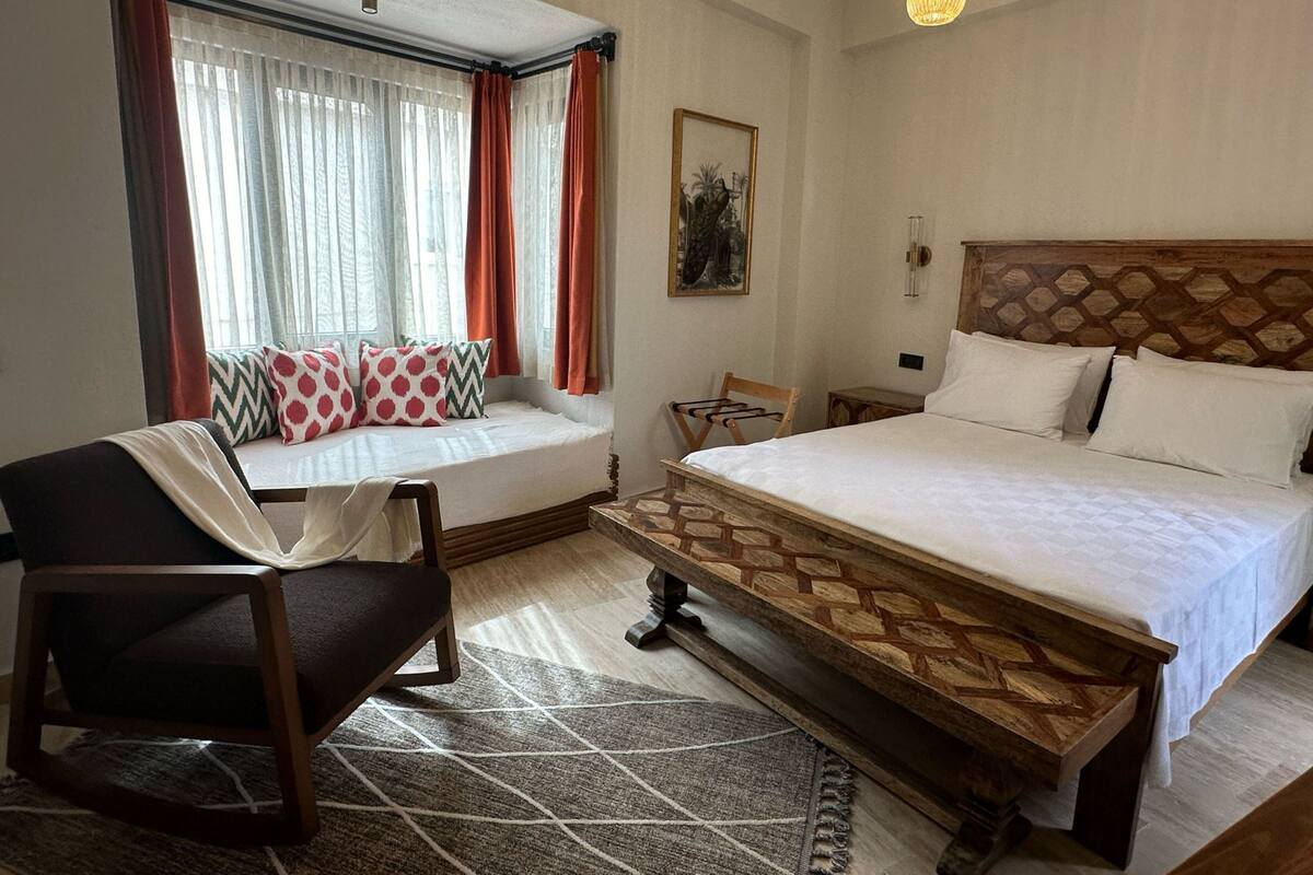 A hotel room with a large bed, a wooden bench, a chair, a window with curtains, and a painting on the wall.