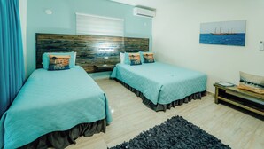 A bedroom with two beds, a wooden headboard, a window with blinds, a painting of a boat on the wall, and a small table with a bag on it. - Comfortable and beautiful Suite (Oranjestad)