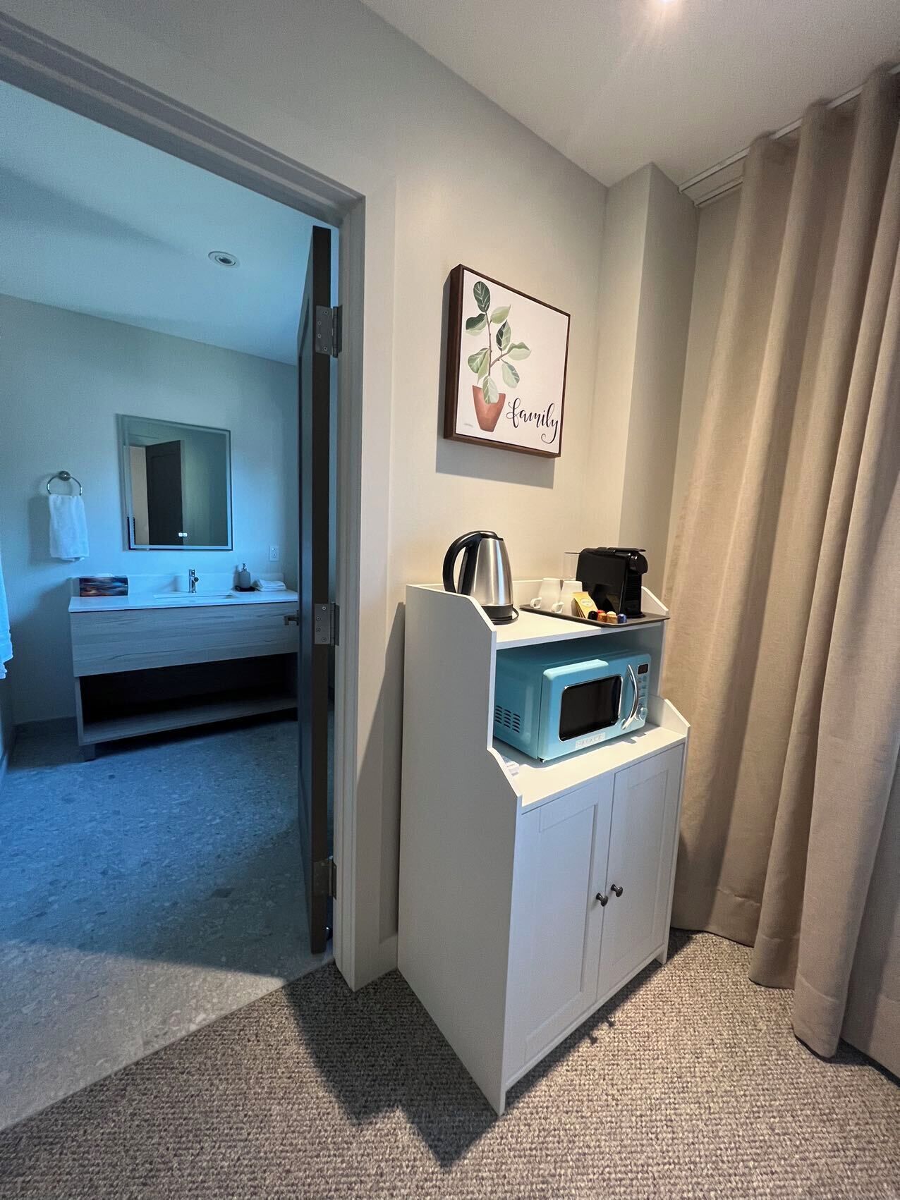 Habitación de hotel con kitchenette, baño con espejo y toallero, y un cuadro en la pared.