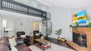 Een ruime woonkamer met een donkerbruine leren bank, een bijzettafeltje en een open haard met bakstenen omlijsting. Een zwart spiraaltrap leidt naar een verdieping boven.