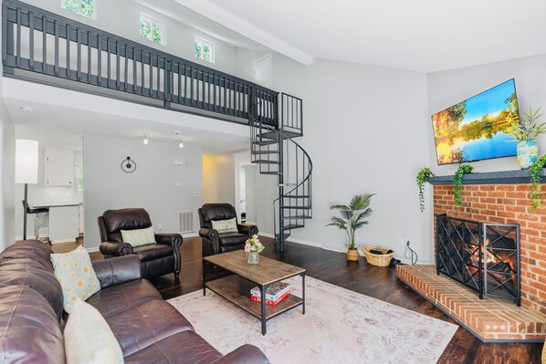 A spacious living area with a dark brown leather sofa, a coffee table, and a fireplace with a brick surround. A black spiral staircase leads to an upper level. - Modern Private Ranch•Very Fast WiFi•Vast Fenced Yard+Big Deck+Fire Pit! (Mint Hill)