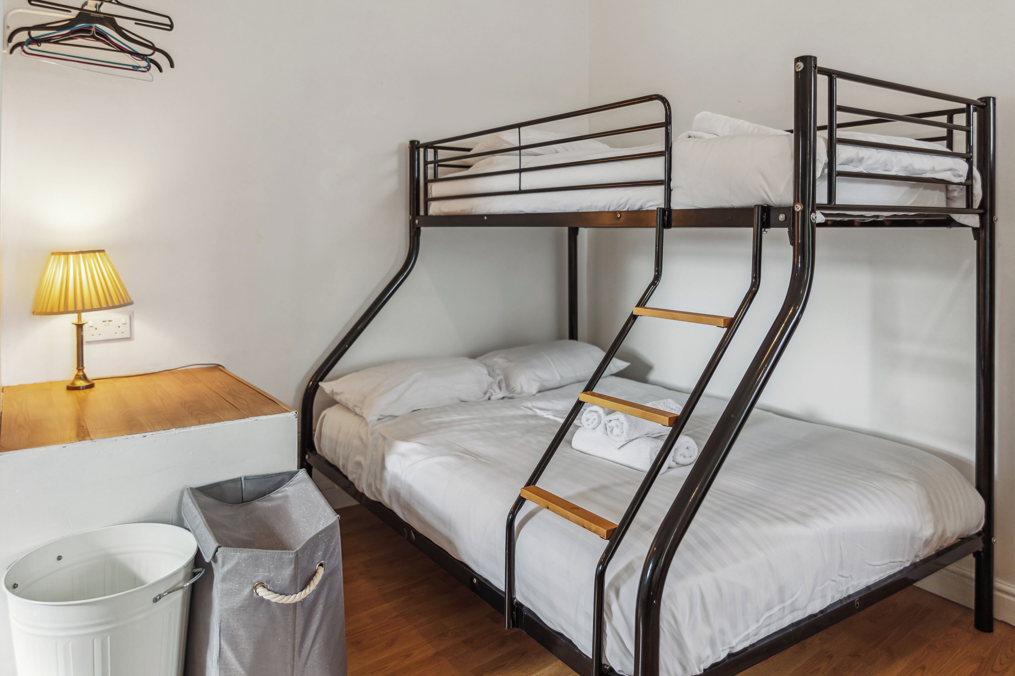 A bunk bed with a wooden ladder, a white trash bin, a grey storage bag, and a lamp on a wooden table.