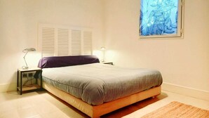 A bedroom with a bed, two bedside tables, a window with a blue artwork, and a white wall. - Pool and seaside apartment  (Cannes)
