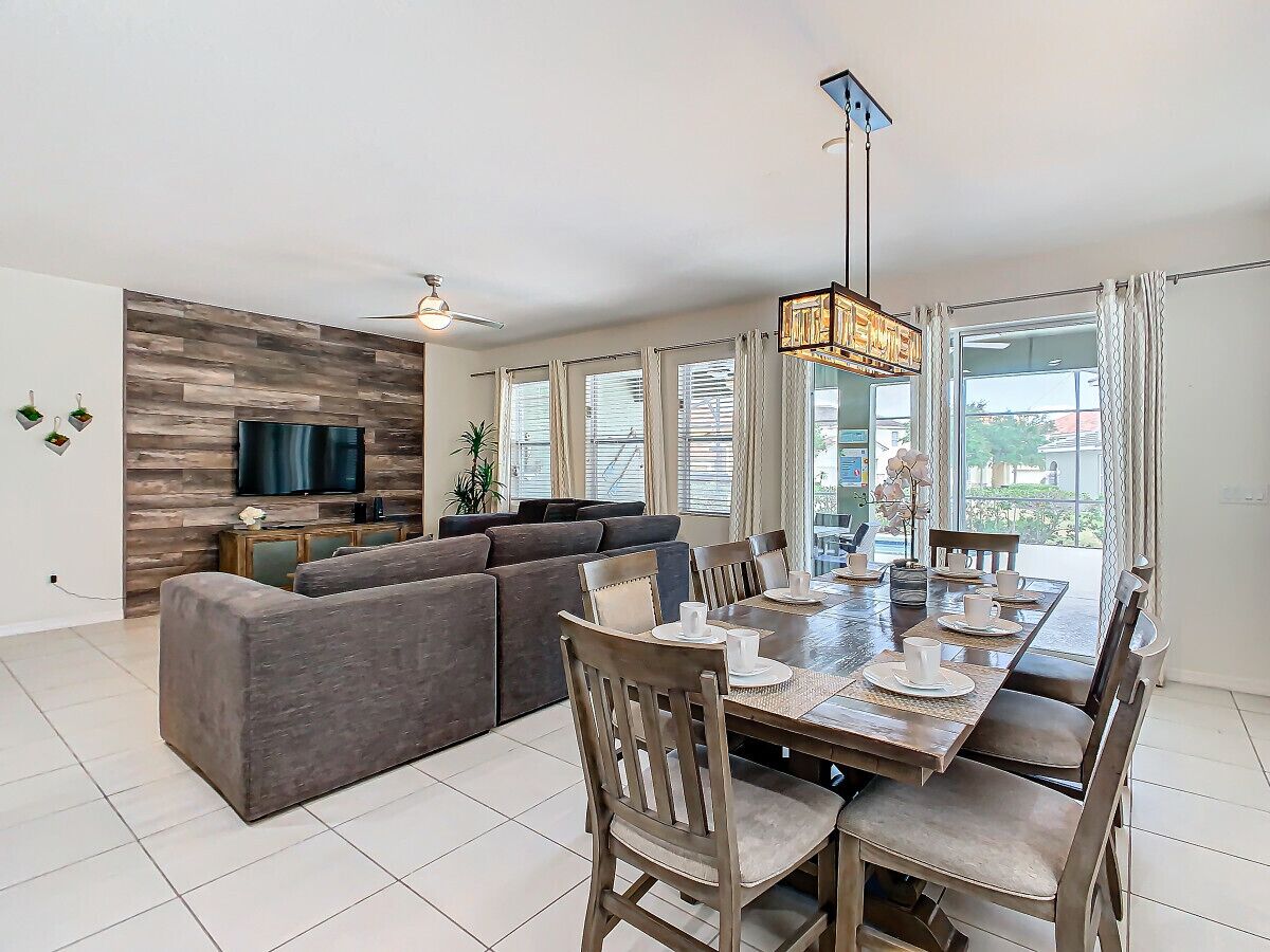 A modern living room with a sectional sofa, a dining table set for four, and a flat-screen TV mounted on a wooden wall.