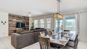 A modern living room with a sectional sofa, a dining table set for four, and a flat-screen TV mounted on a wooden wall. - Serene 7-Bedroom Villa in Solterra with Pool (Davenport)