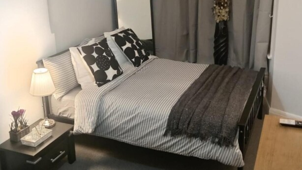 Un letto rifatto con lenzuola a righe e una coperta grigia, una lampada da comodino e un piccolo tavolino con una pianta e un oggetto decorativo.