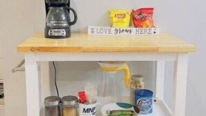 Un chariot de cuisine blanc, doté d’une étagère supérieure en bois où sont posés une machine à café, des chips et un panneau sur lequel on peut lire : « L’AMOUR s’épanouit ICI ». Le chariot comporte deux étagères en dessous, contenant des bocaux, une tasse et une assiette avec de la nourriture.