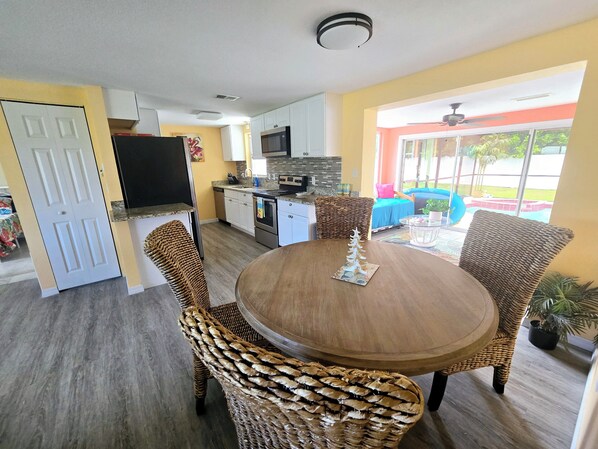 A dining area with wicker chairs and a round table, a kitchen with white cabinets and a microwave, and a view of a pool through large windows.
