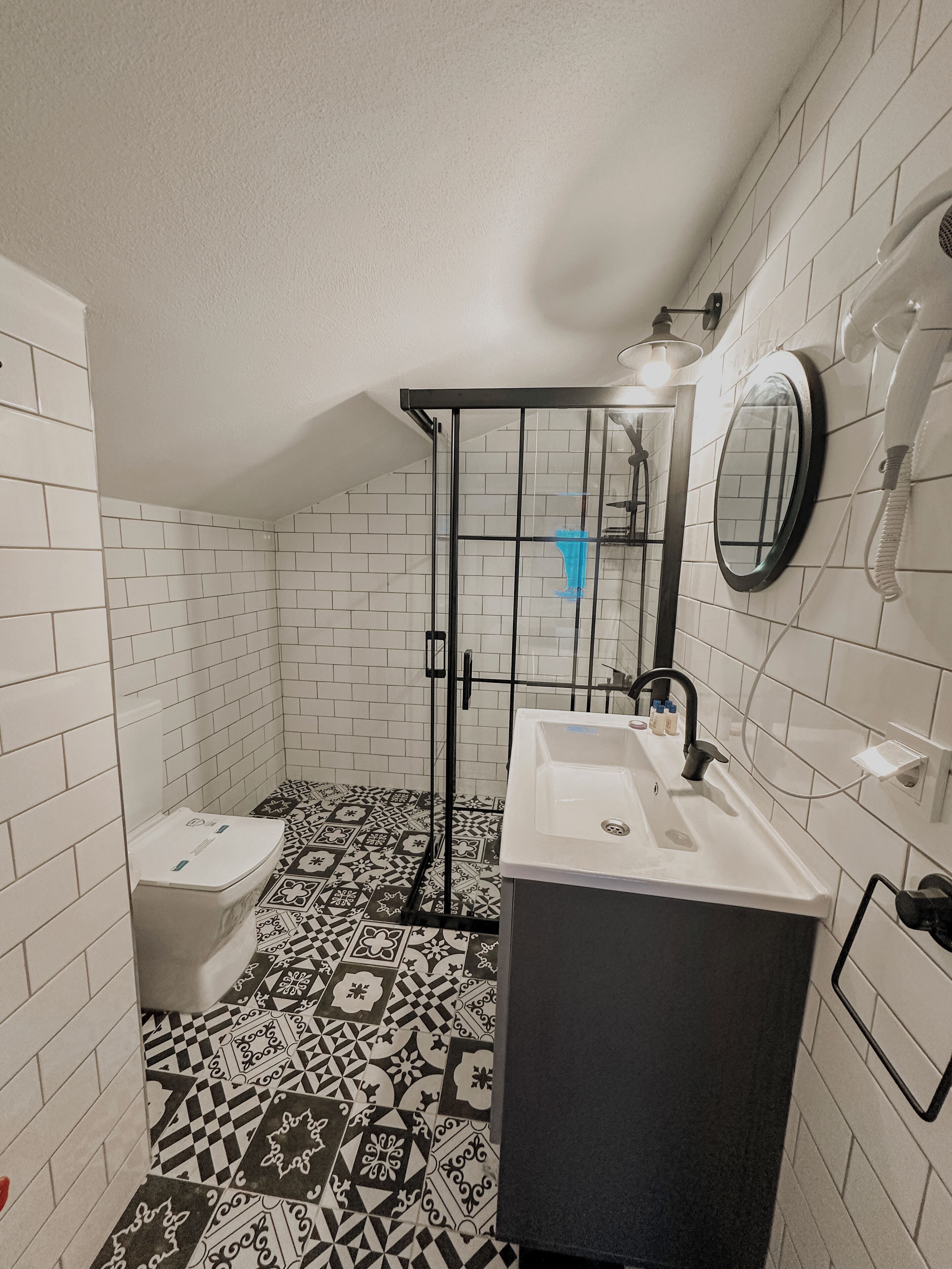 A bathroom with white tiled walls, a black and white patterned floor, a shower enclosure, a toilet, and a sink with a mirror.