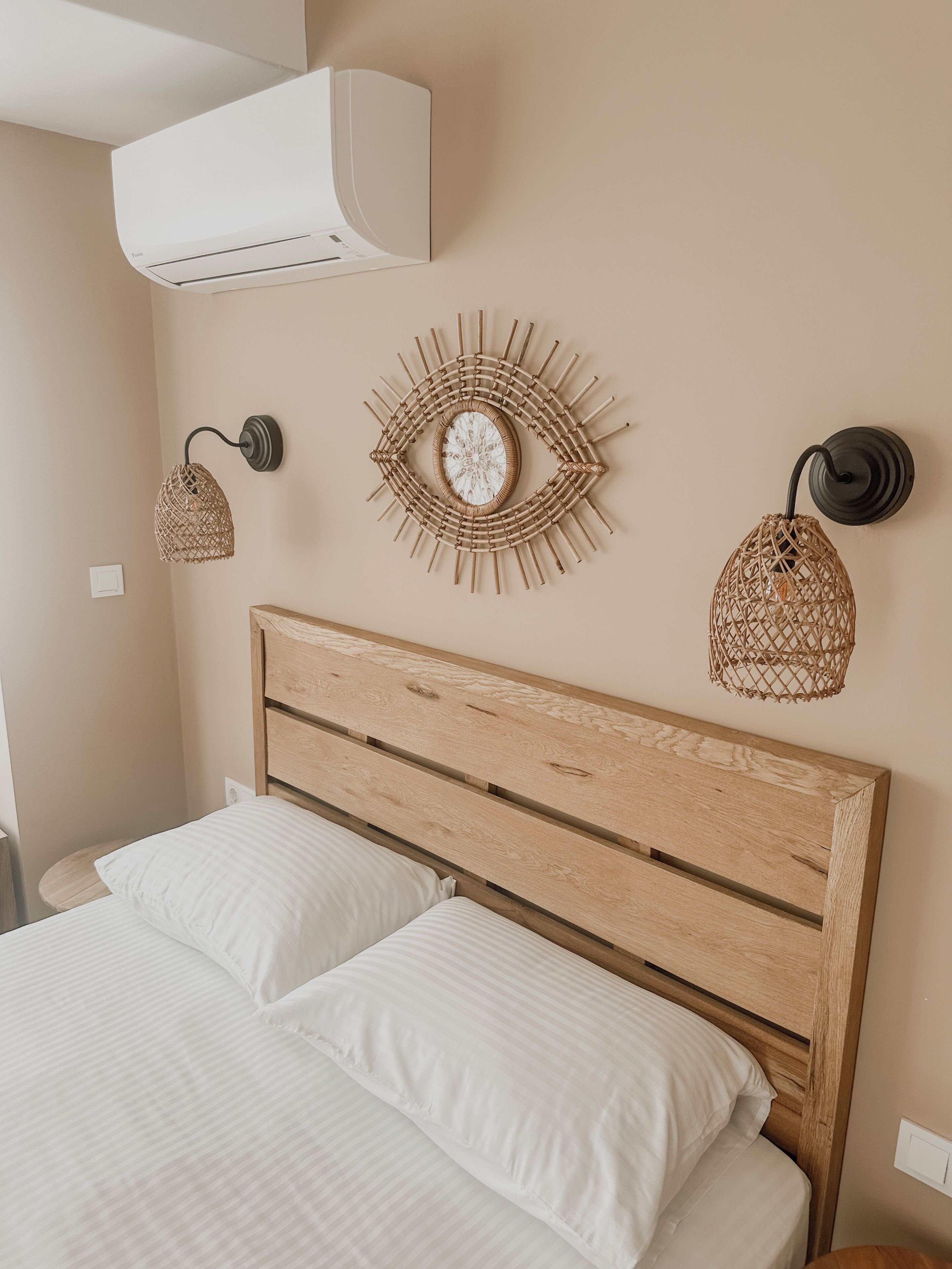 Ein Schlafzimmer mit einem Holzkopfende, zwei weißen Kissen, einer Wand-Klimaanlage und zwei Wandleuchten mit gewobenen Lampenschirmen.