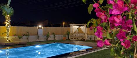 Una piscina con césped artificial, una palmera y flores rosadas.